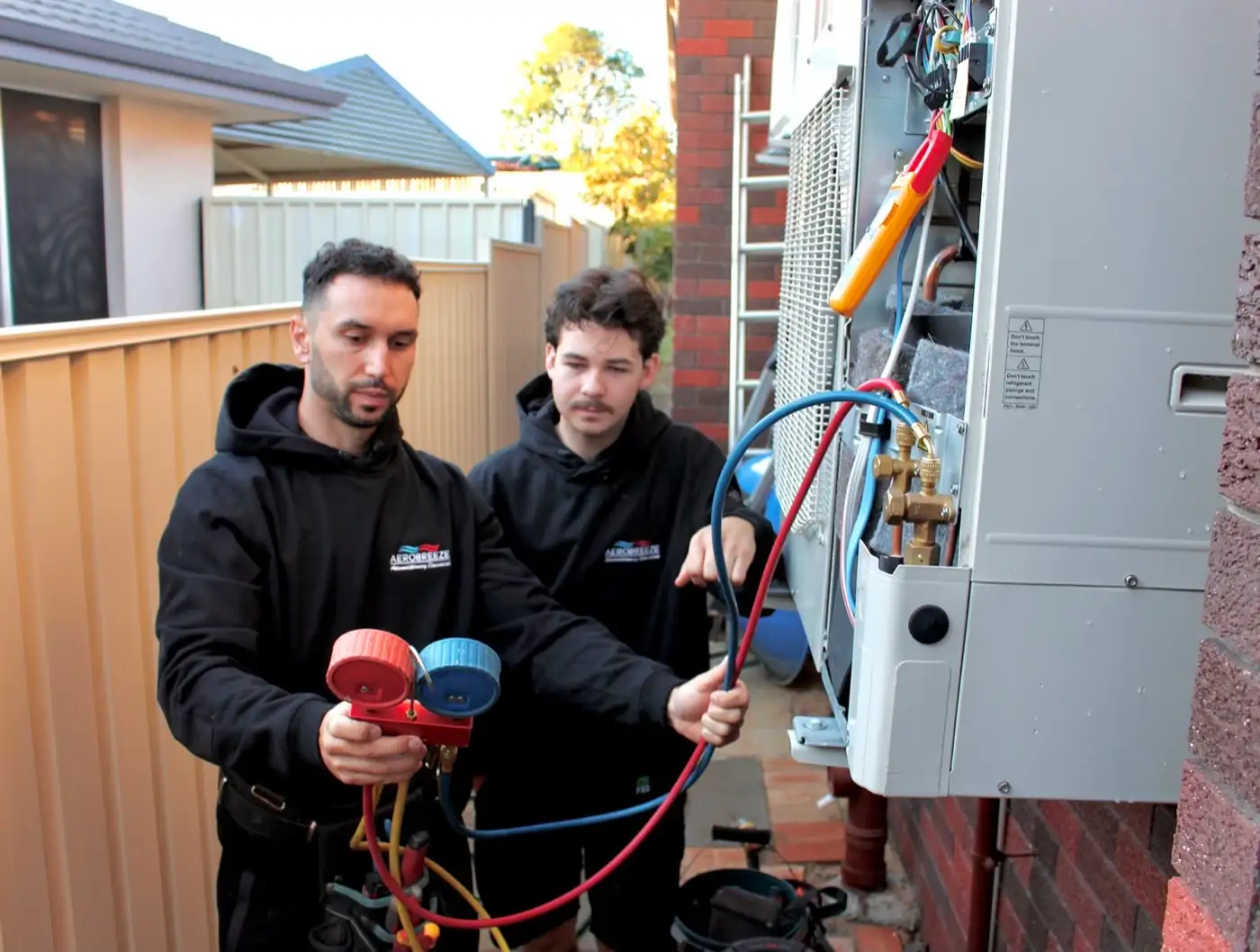 Two Aerobreeze technicians reading manifold gauges at an opened outdoor unit, checking the refrigerant charge