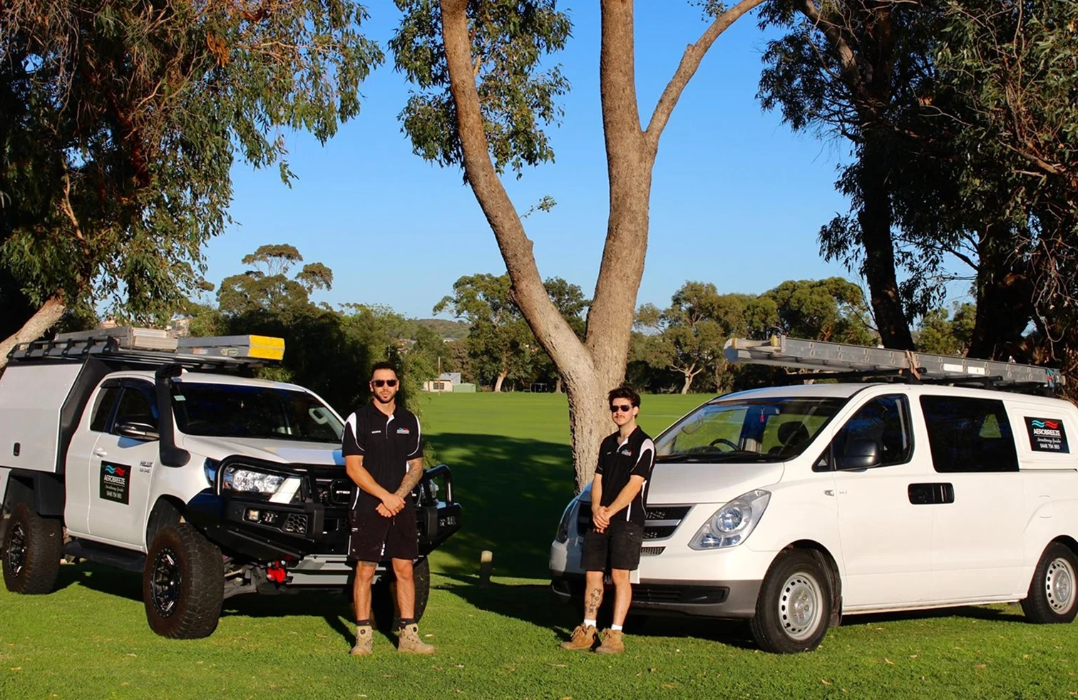 Two Aerobreeze technicians standing in front of the marked ute and van, parked on grass under a gum tree